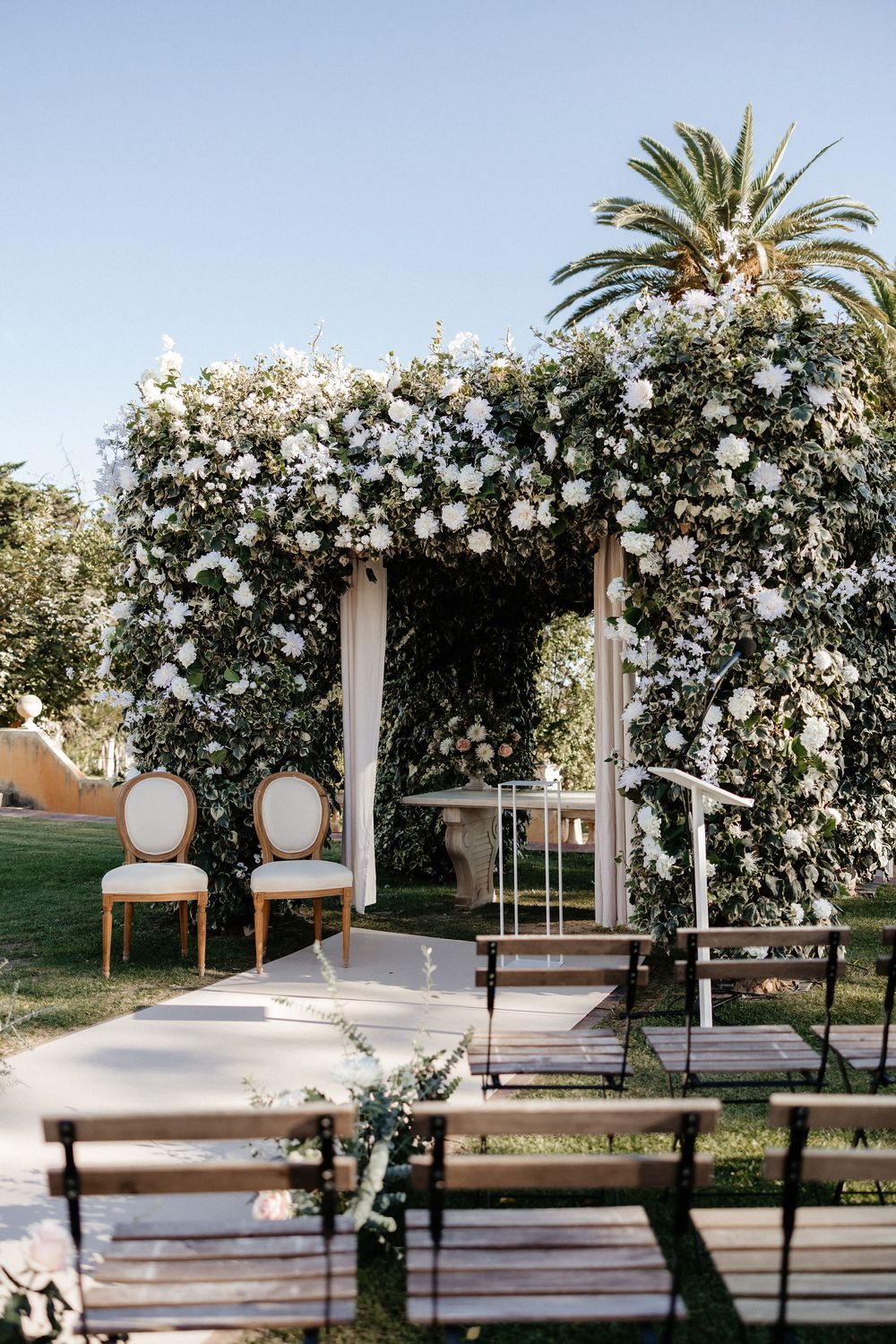 Elegant outdoor wedding ceremony setup with white floral arch and wooden benches arranged in rows under palm trees.