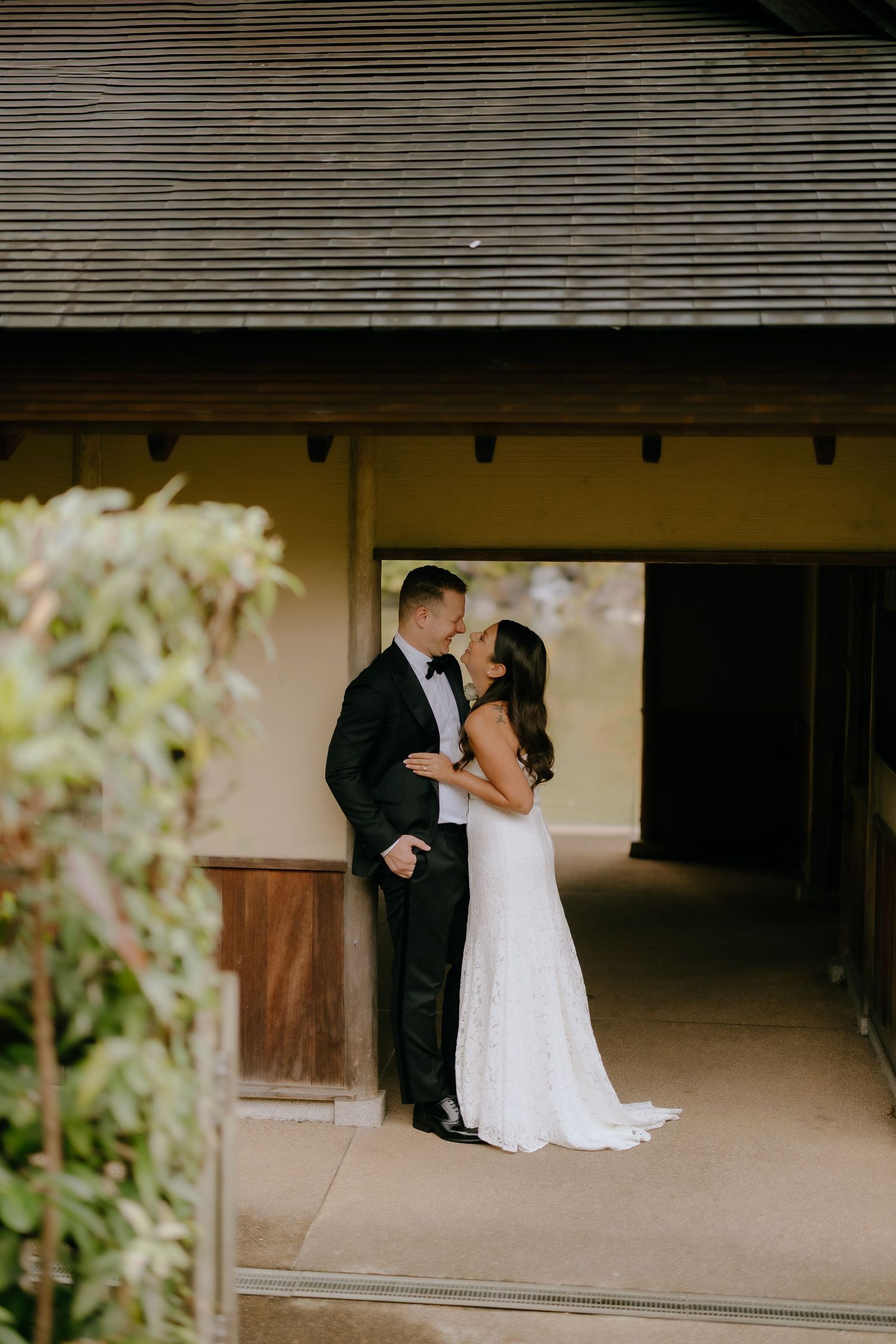 A bride and groom share an intimate moment in a covered walkway with white flowers blooming nearby.