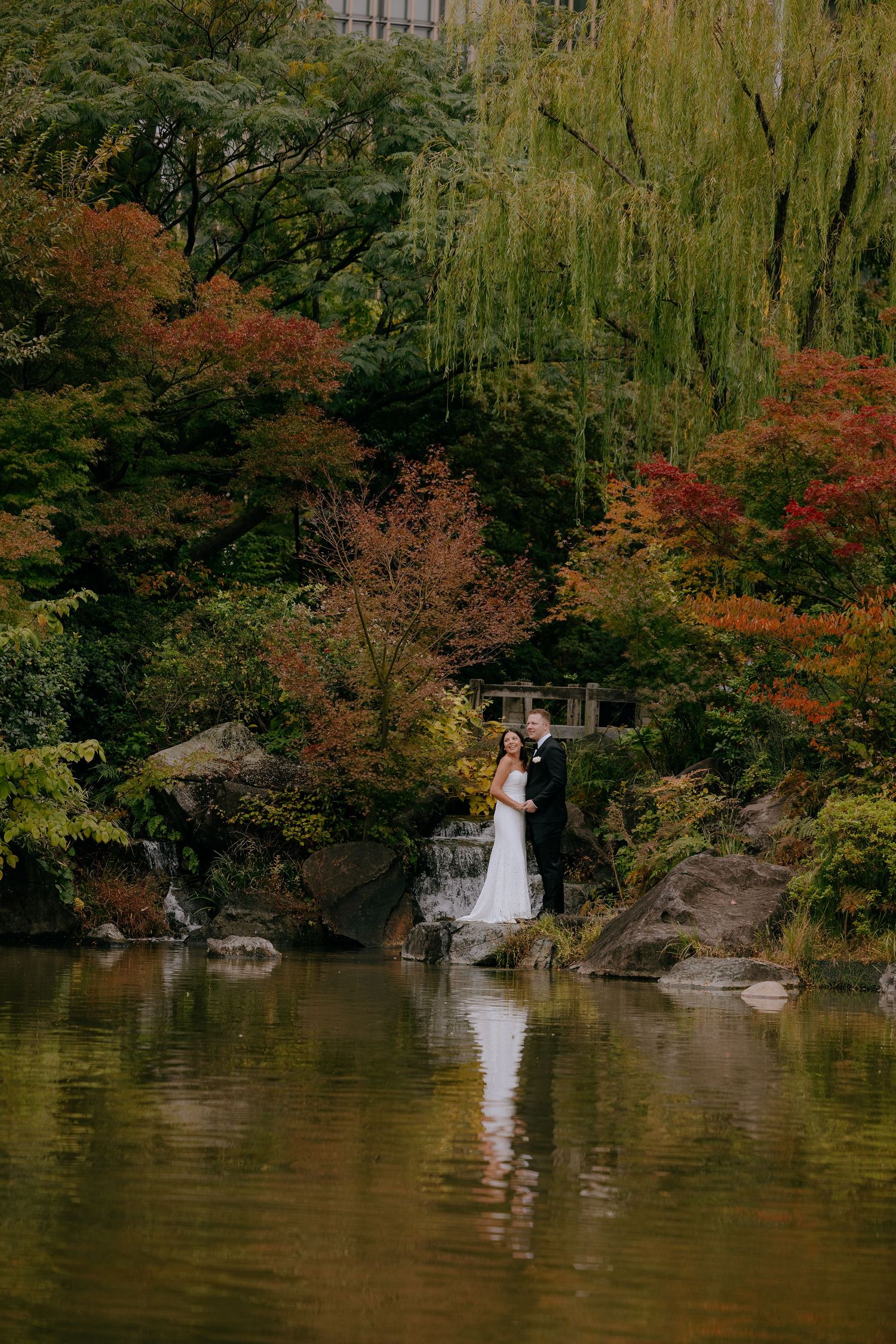 A couple in wedding attire stands by a tranquil pond surrounded by autumn trees with vibrant red and orange foliage.