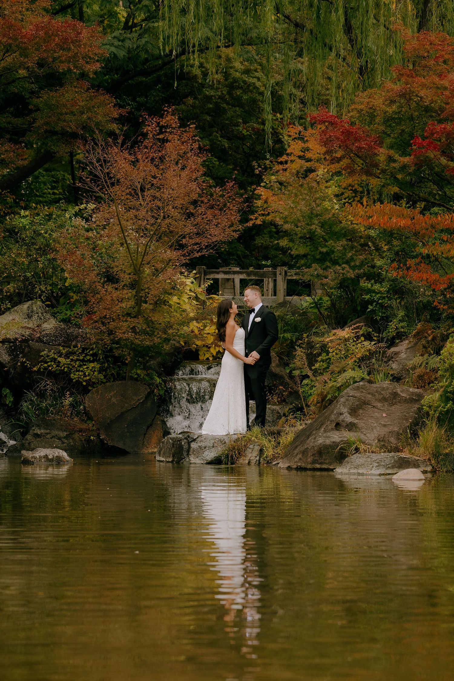 A couple in wedding attire stands by a tranquil lake surrounded by autumn foliage and rocky outcrops.
