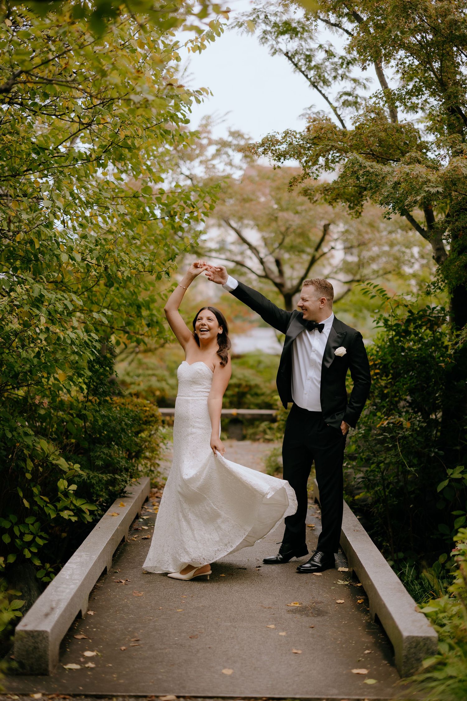 A couple in formal wedding attire dances playfully on a garden path surrounded by autumn foliage.