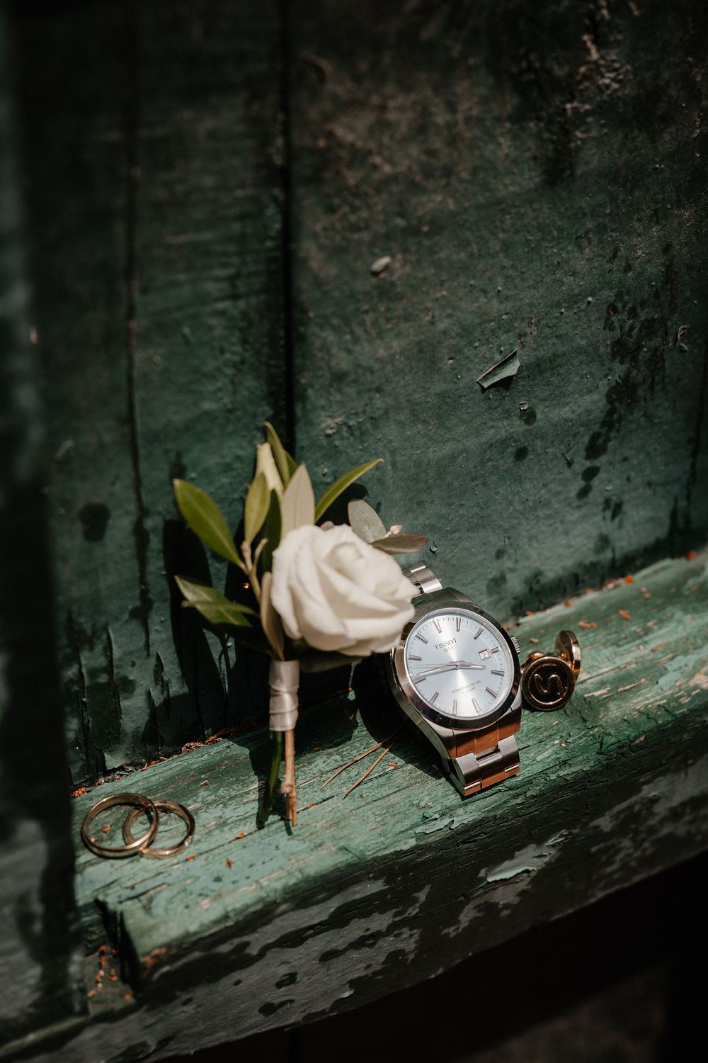 Wedding details including gold rings, white rose, vintage pocket watch, and bottle arranged on weathered green wood surface.