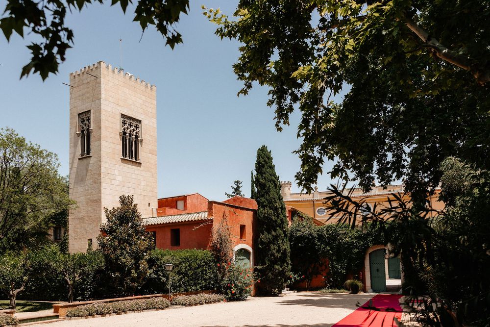 A stone tower and red building with Mediterranean architecture set against a blue sky with a red carpet walkway.
