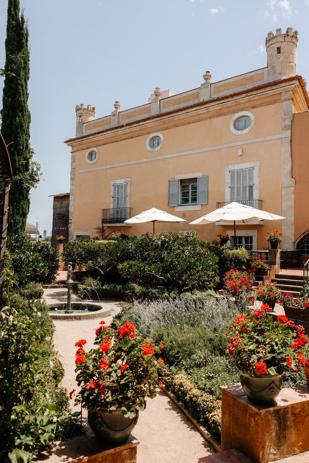 Mediterranean villa with peach facade, ornate towers, and garden terrace featuring red flowers and white umbrellas.