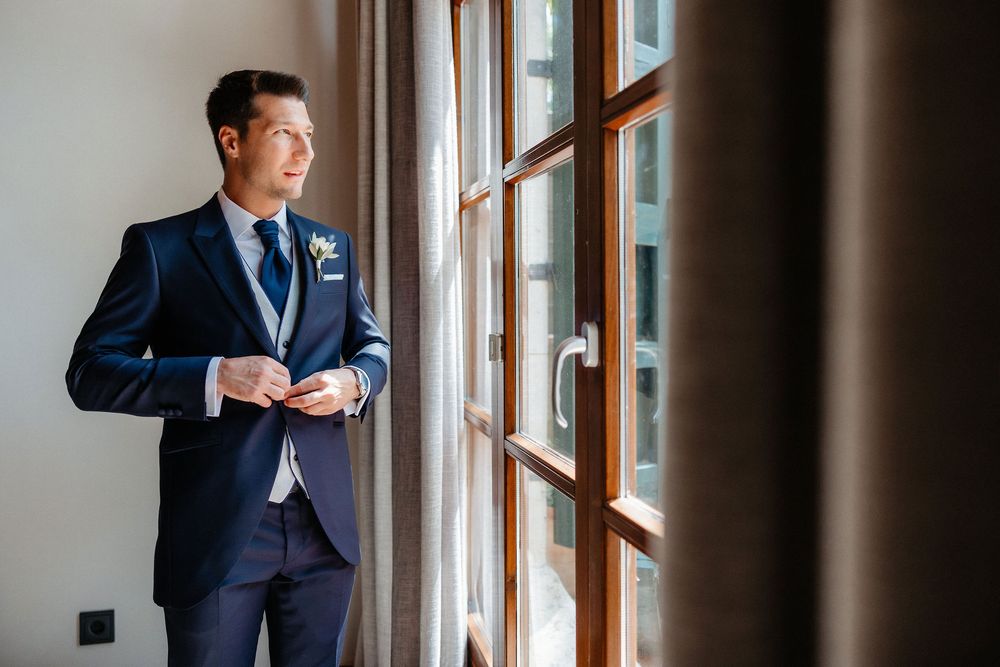 Groom in navy suit adjusts sleeve while standing by window in elegant getting ready moment.