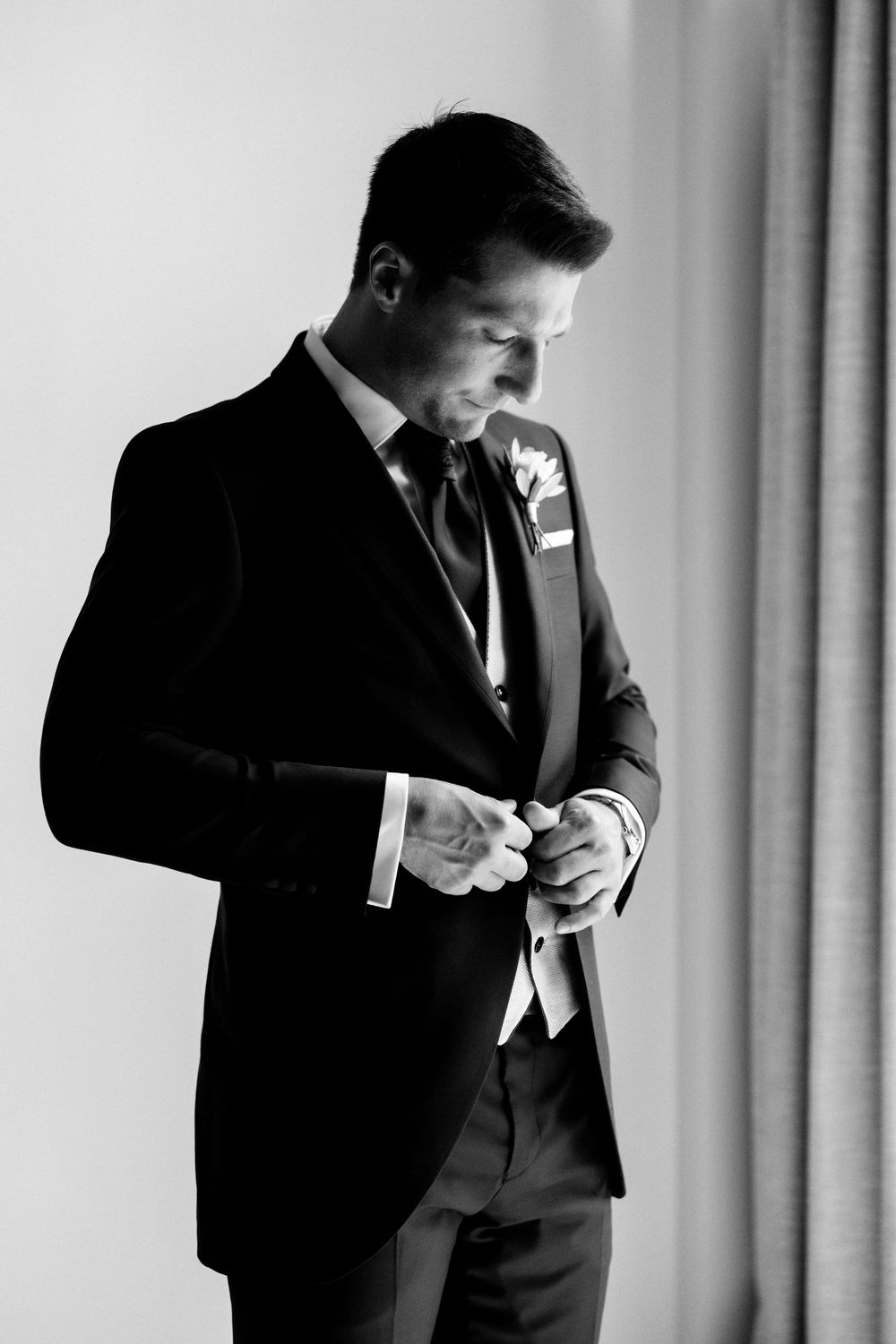 Black and white photo of a groom adjusting his tie while getting ready for a wedding ceremony.