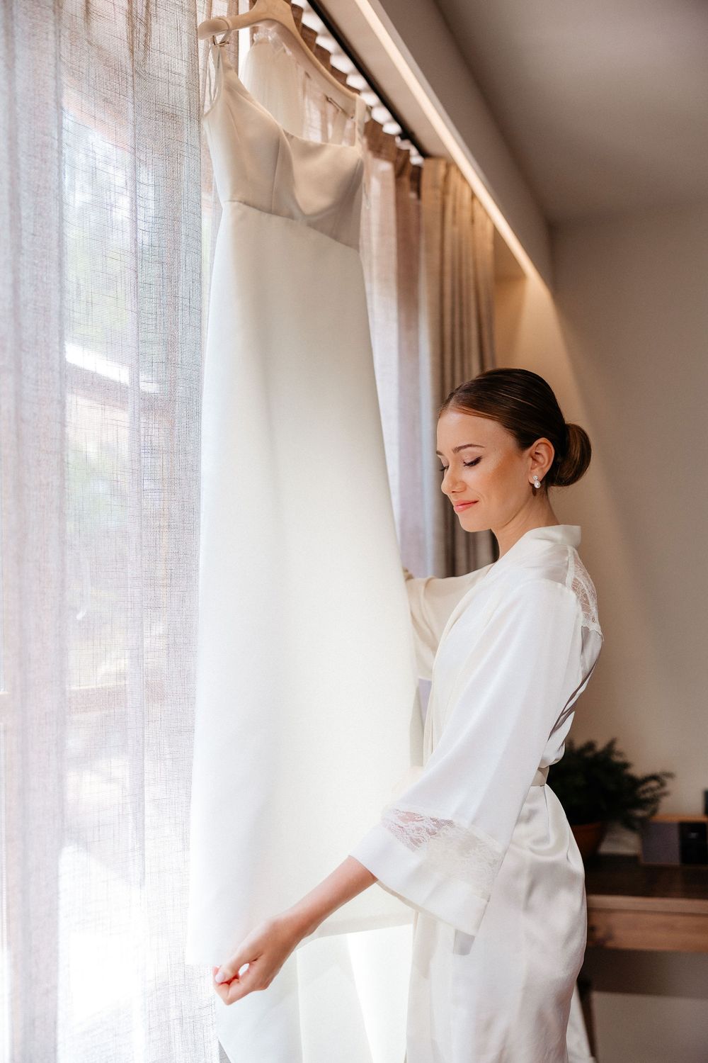 Bride in white robe preparing for wedding day near sunlit windows in elegant getting ready room.
