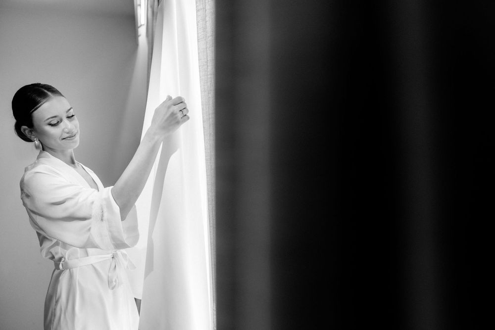Bride in white robe stands gracefully by window during wedding day preparations.