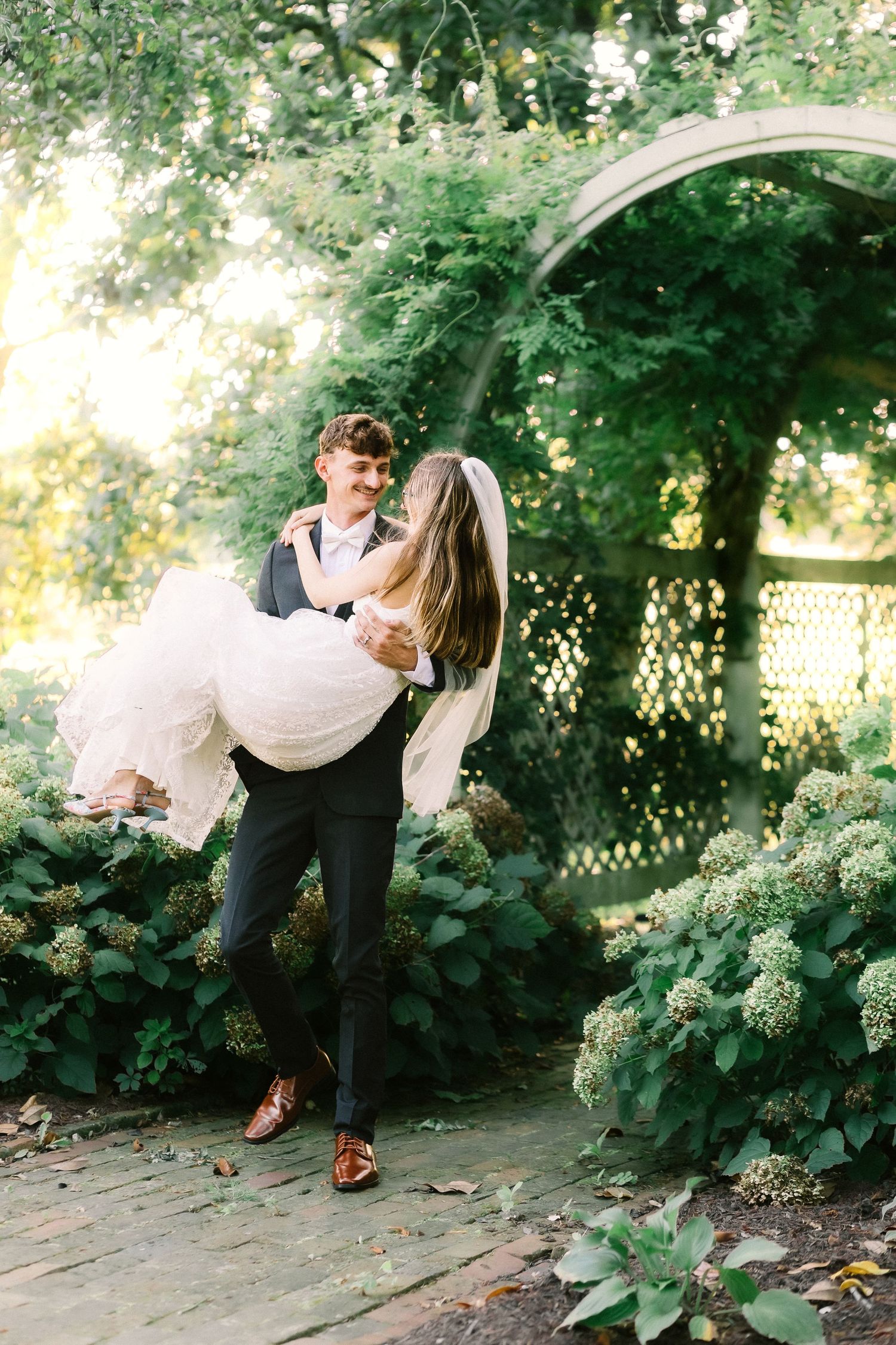 A joyful wedding moment captured outdoors with the groom in a dark suit and pink bowtie standing with his bride in white.
