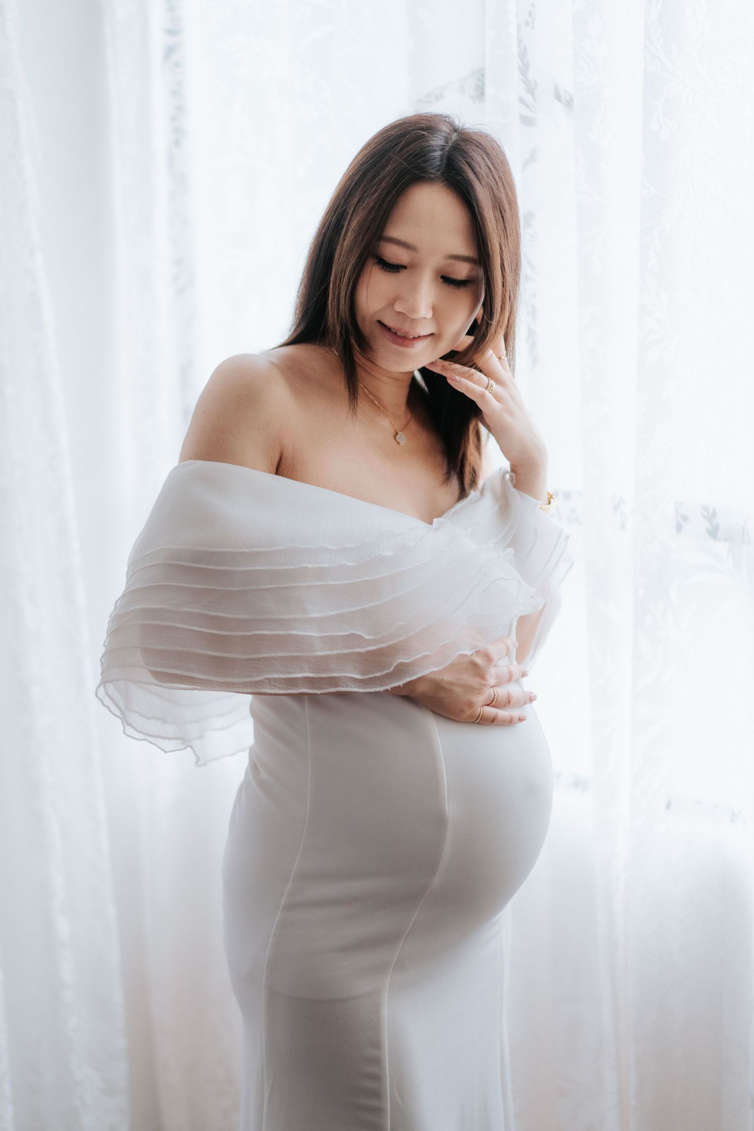 A pregnant woman in a white flowing maternity dress poses gracefully by sheer curtains in soft natural lighting.
