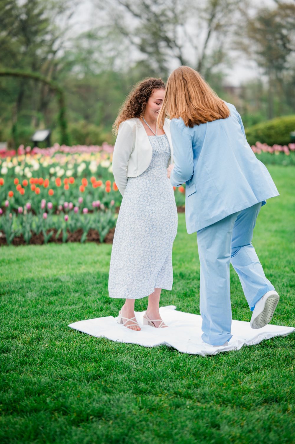 A couple in white and blue outfits walking together on a green lawn with tulips in the background.