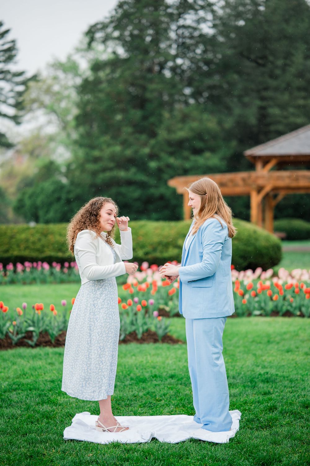 Two people stand together on a white cloth surrounded by blooming tulip gardens and a wooden gazebo.