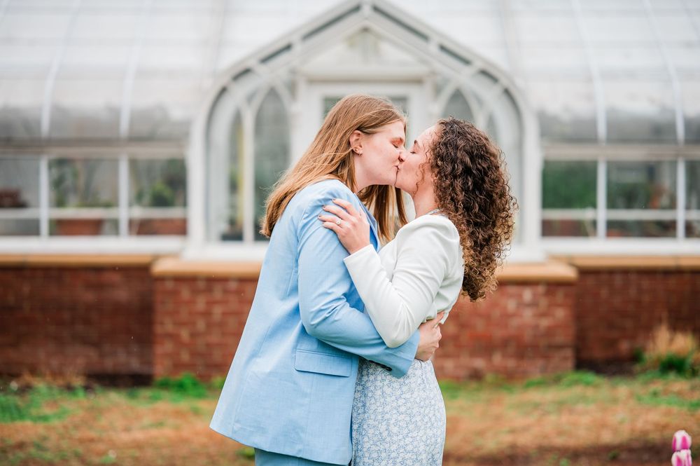 A couple shares a tender kiss in front of a brick building with glass windows on a bright day.