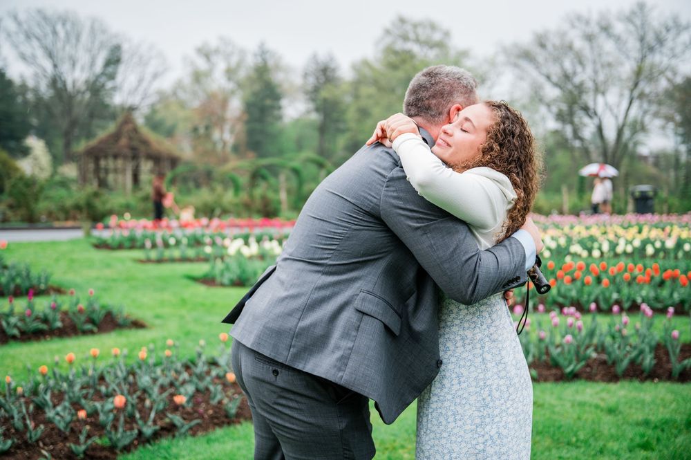 A couple embraces in a garden setting with colorful tulip beds and a gazebo in the background.