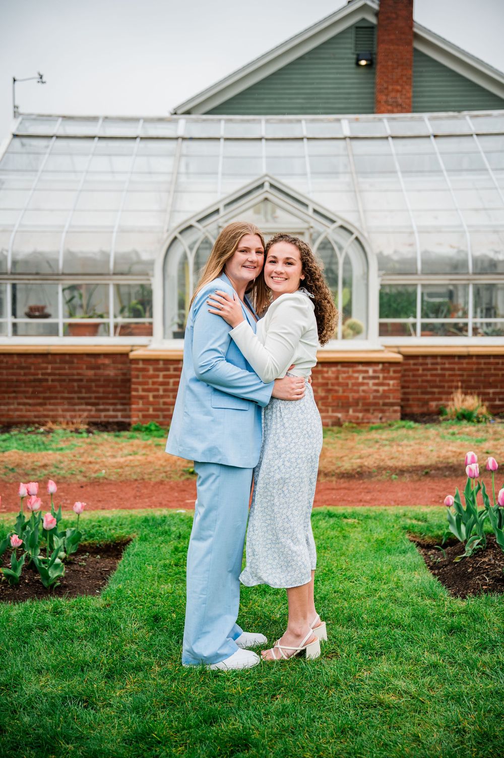 A tender embrace captured in front of a historic greenhouse surrounded by spring flowers.