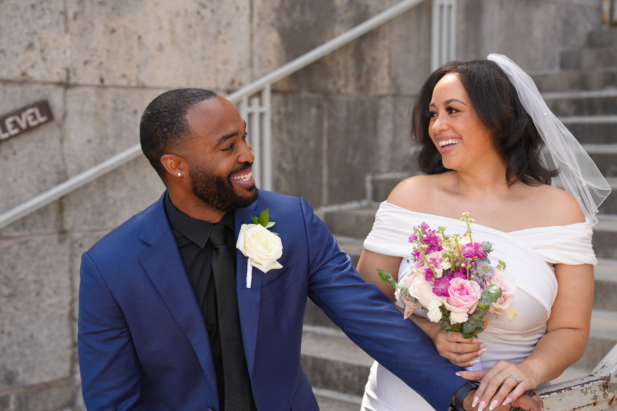 Happy newlywed couple in wedding attire smiling at each other on outdoor steps.