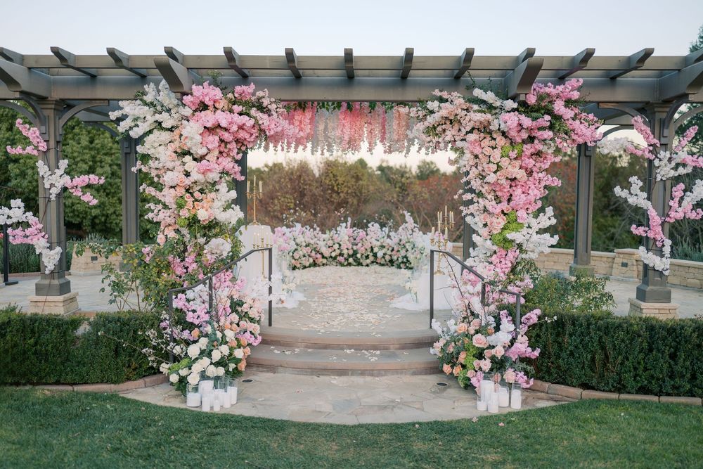 Elegant outdoor wedding arch decorated with pink cherry blossoms and white flowers under a wooden pergola in a garden setting.