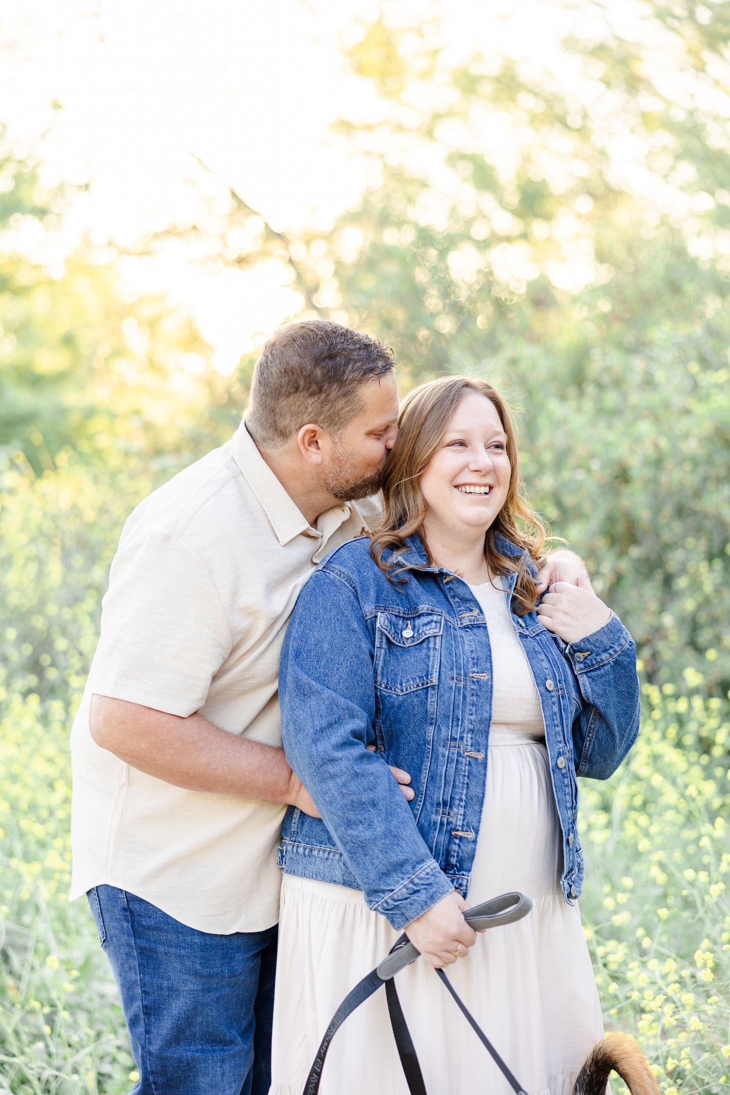 Couple shares playful moment outdoors in denim jacket and white shirt against natural greenery background.