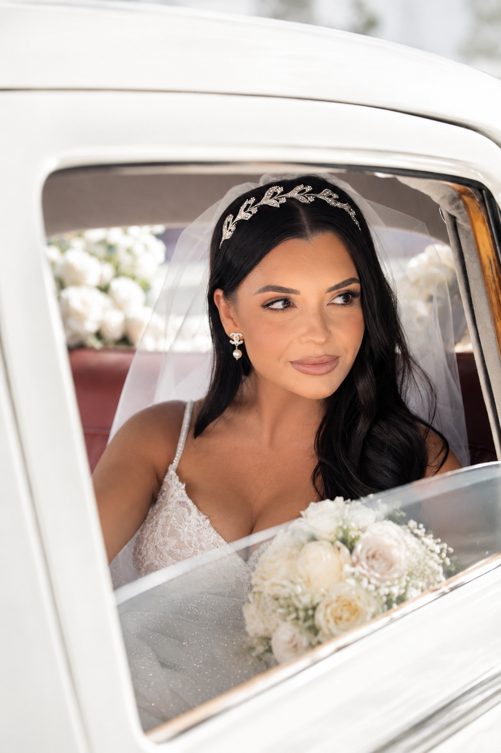 Beautiful dark-haired bride in lace gown with crystal headband and veil holding white bouquet in vintage car.