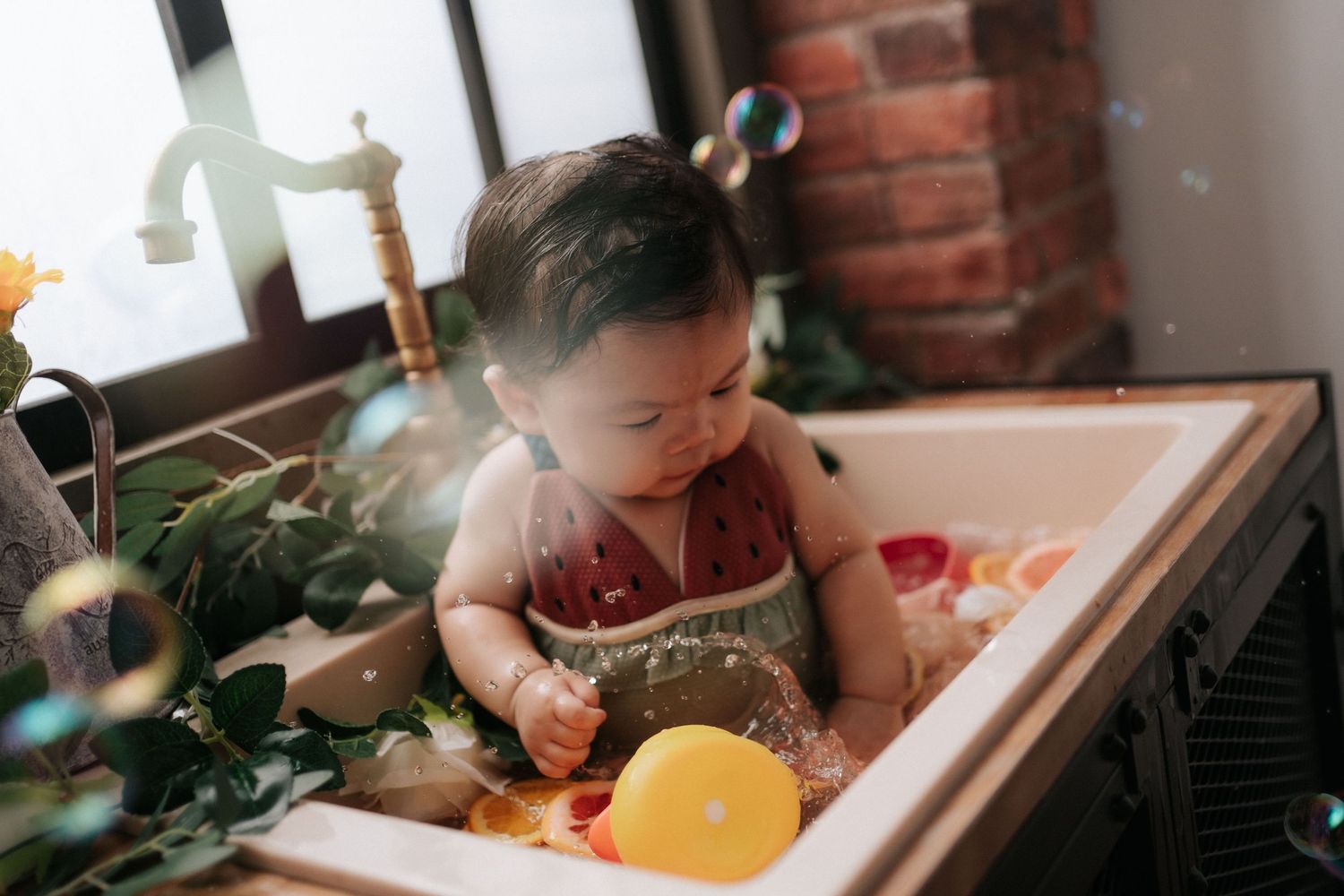 Small child plays in kitchen sink with yellow balloon and greenery while soft natural light streams through window.