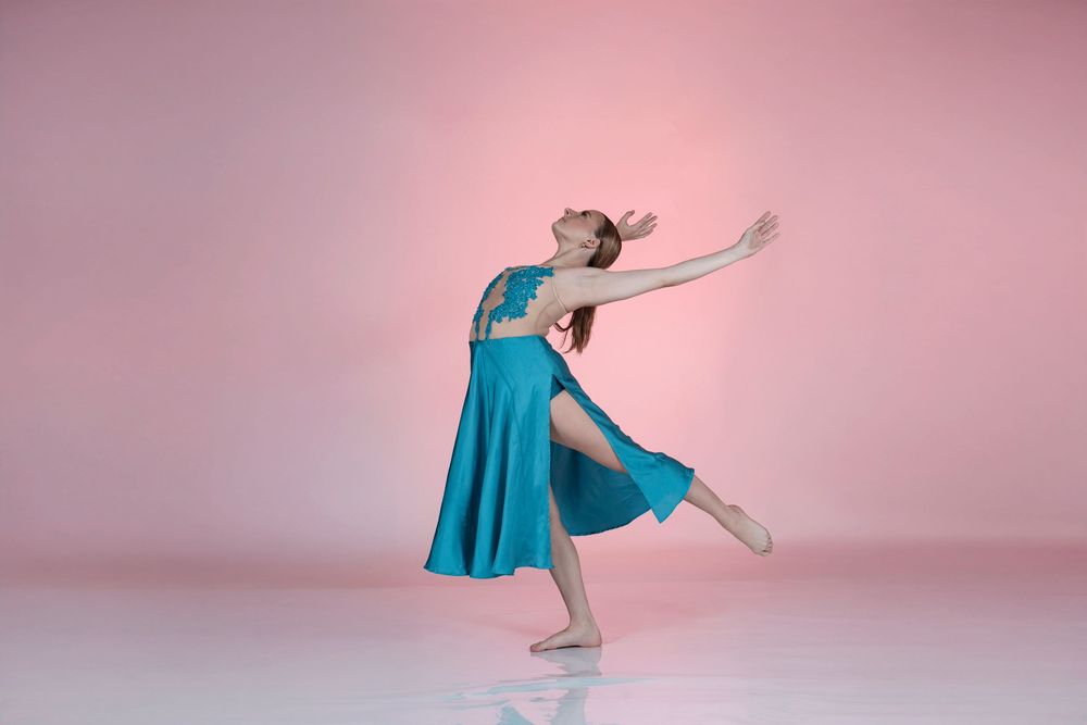 Dancer in flowing turquoise dress executes elegant movements against pink backdrop.