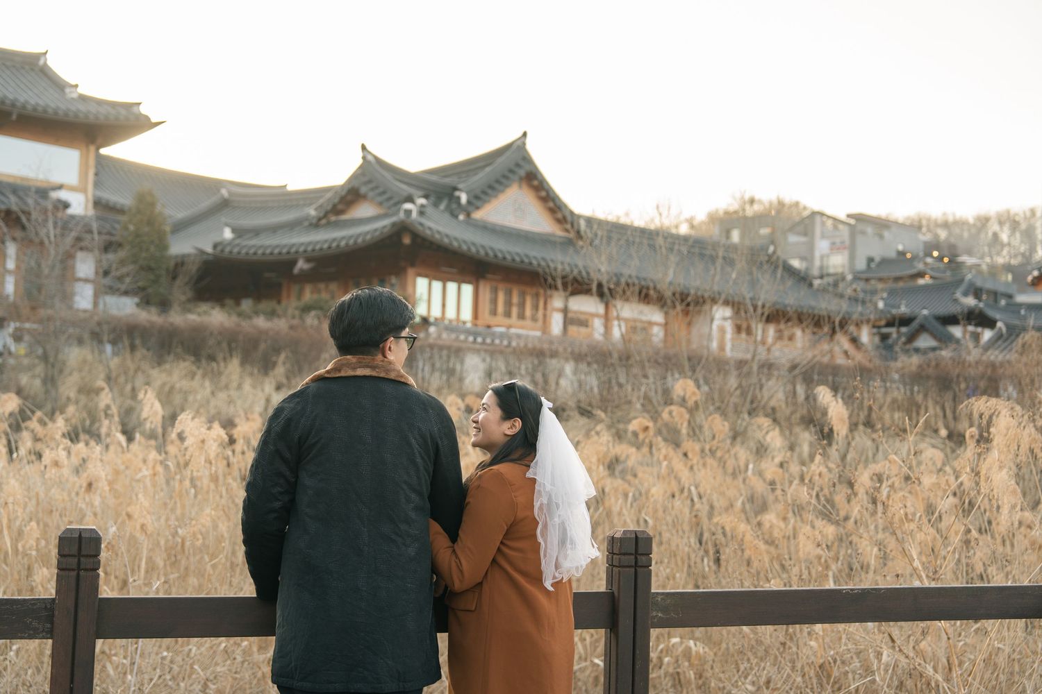 A couple in winter coats stand together on a wooden deck overlooking traditional Asian architecture and mountains.