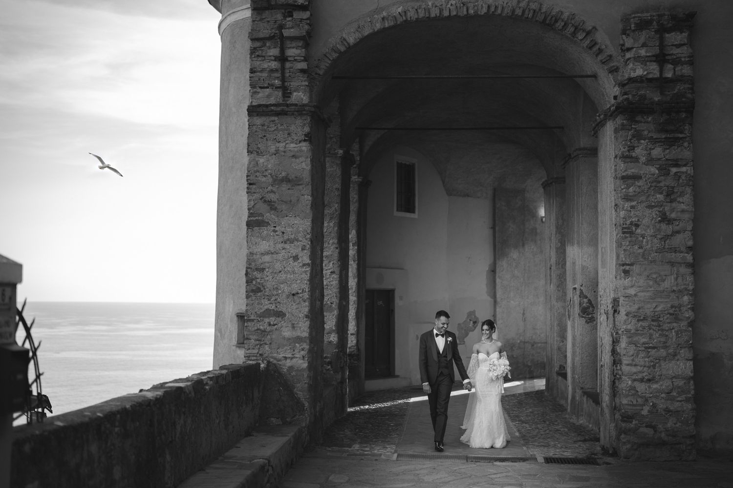 Sposi che camminano mano nella mano sotto arcata storica vista mare in Liguria