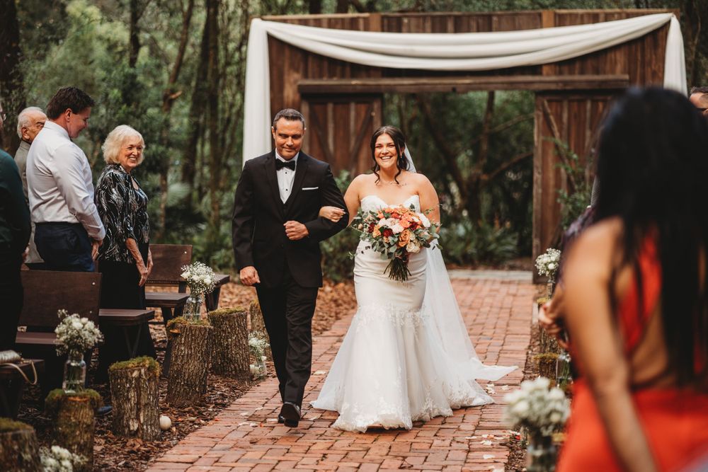 A wedding ceremony processional on a brick pathway with rustic log seating and floral decorations.