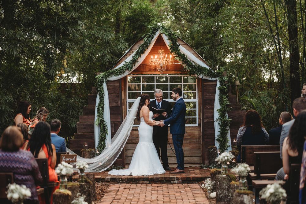 Wedding ceremony under a decorated wooden gazebo with white fabric draping and floral arrangements.