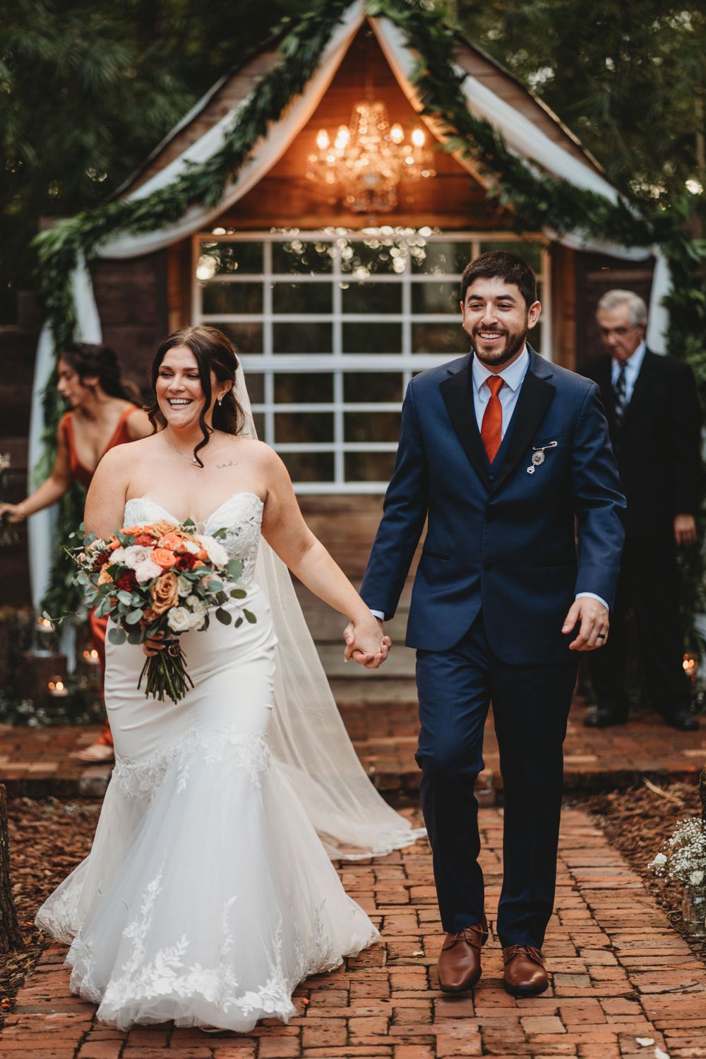 A joyful wedding exit down a brick pathway from a wooden gazebo decorated with lights and greenery.