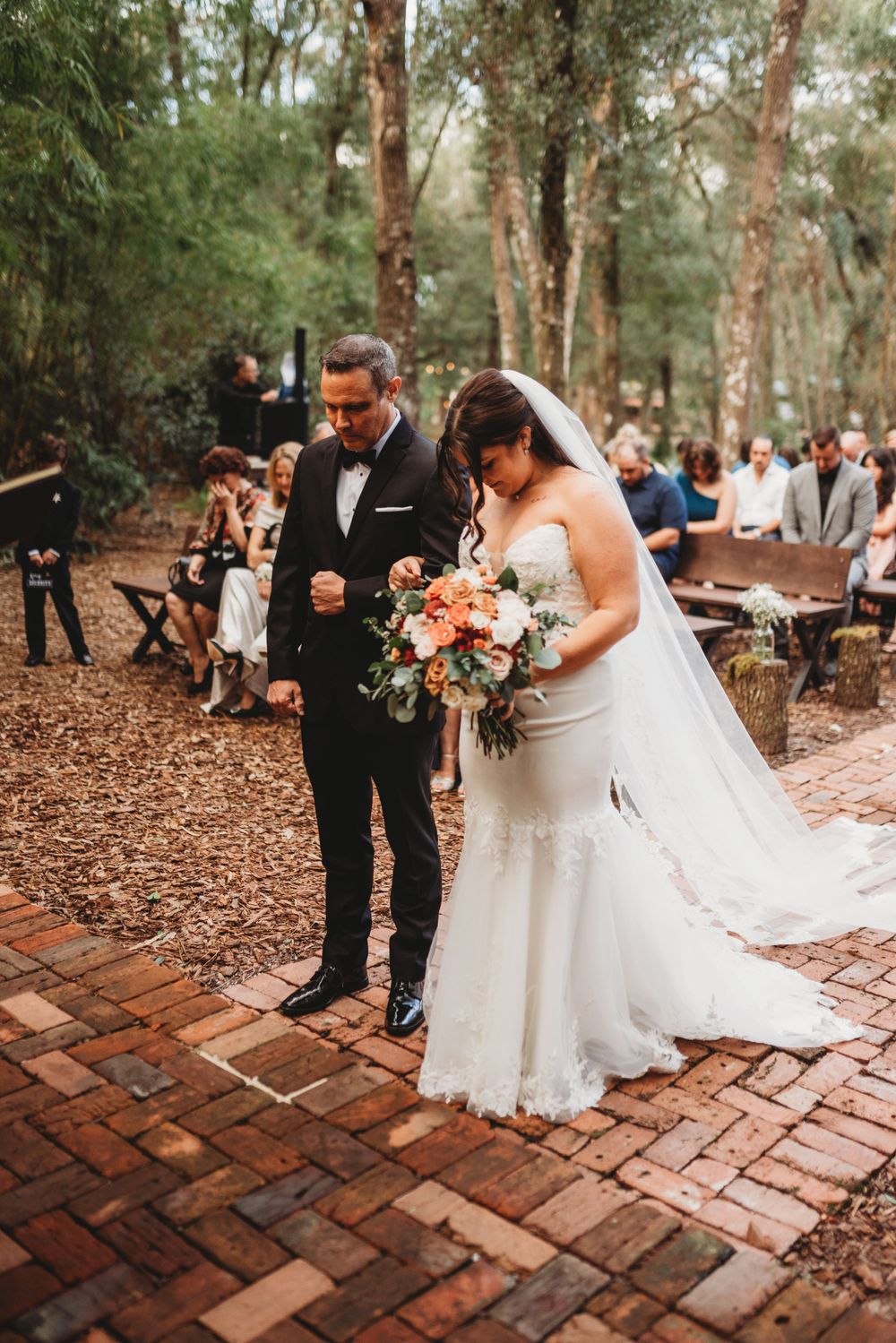 A wedding couple walks down a brick path surrounded by tall trees in an outdoor forest ceremony.