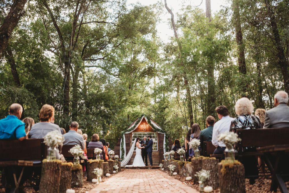 A rustic outdoor wedding ceremony with guests seated on wooden benches and hay bales along a forest path.