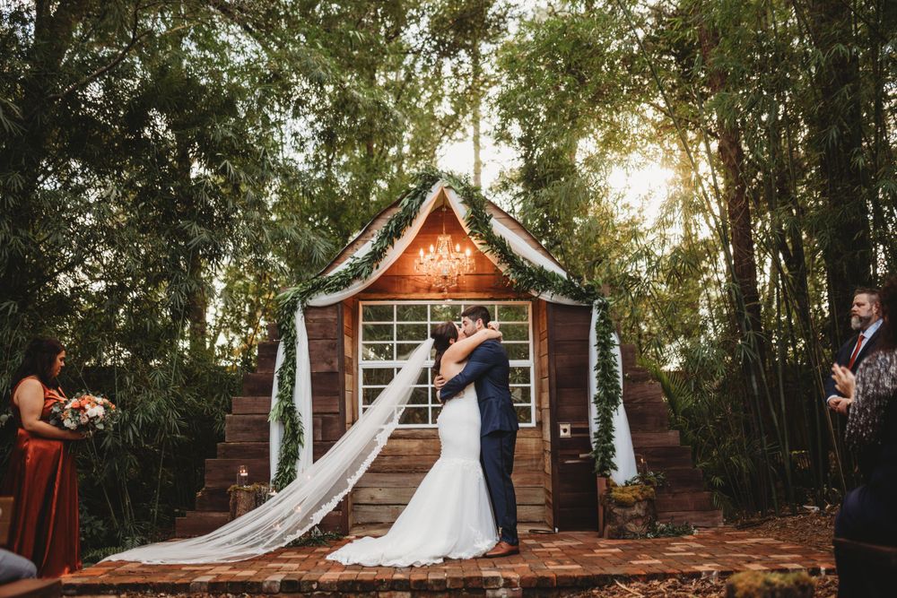 A magical forest wedding ceremony under a wooden arch decorated with string lights at dusk.