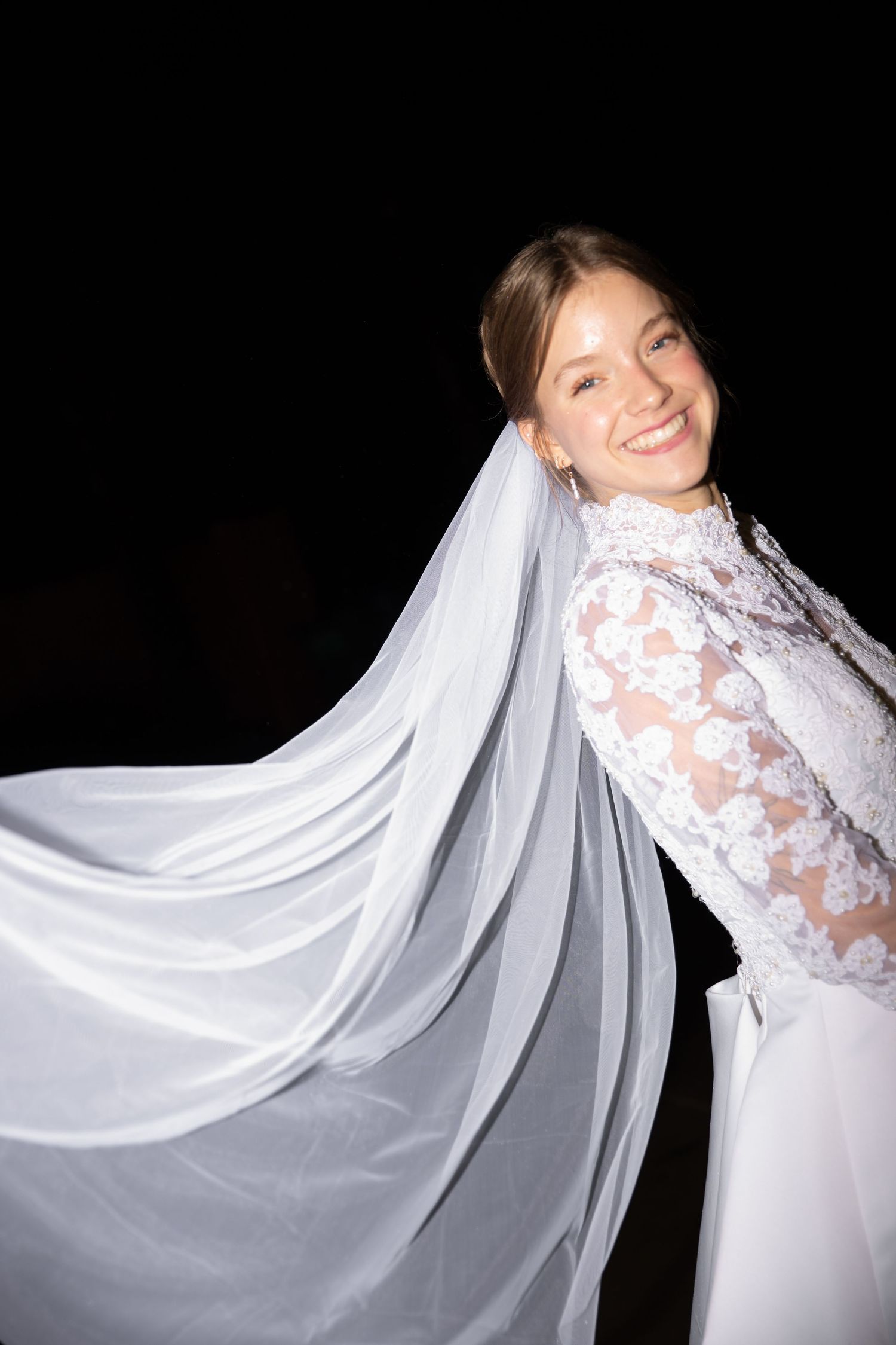 Bride in long sleeve lace wedding dress with flowing veil twirling against dark background.