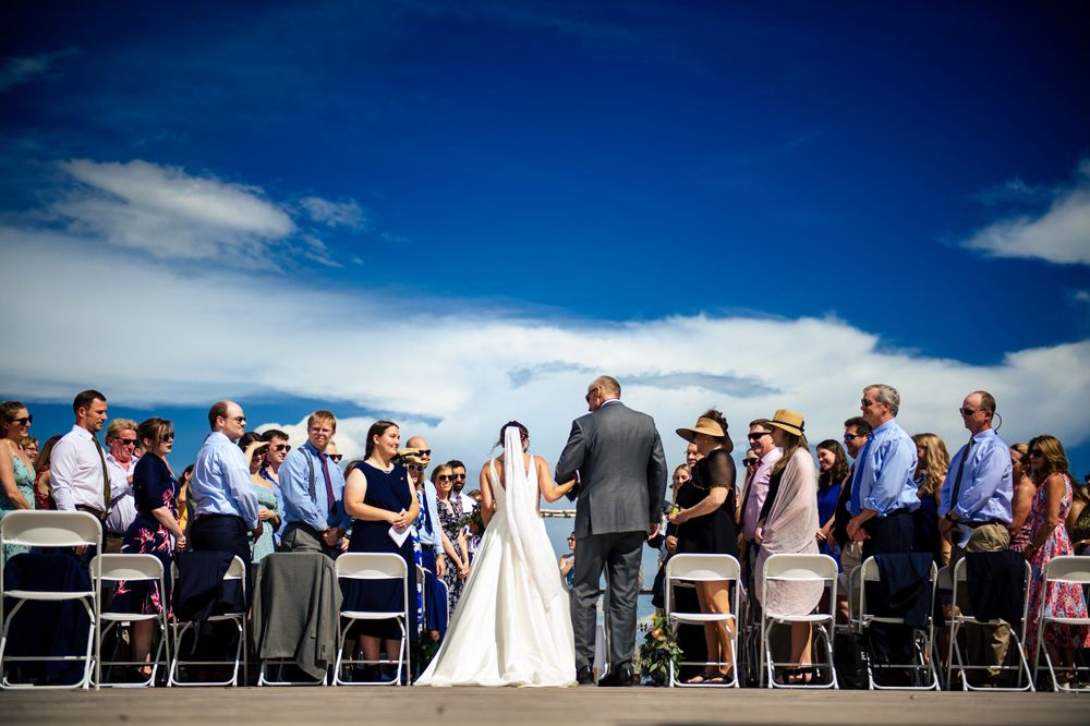 Bride and groom exchange vows at outdoor wedding ceremony under blue sky with seated guests in white chairs.