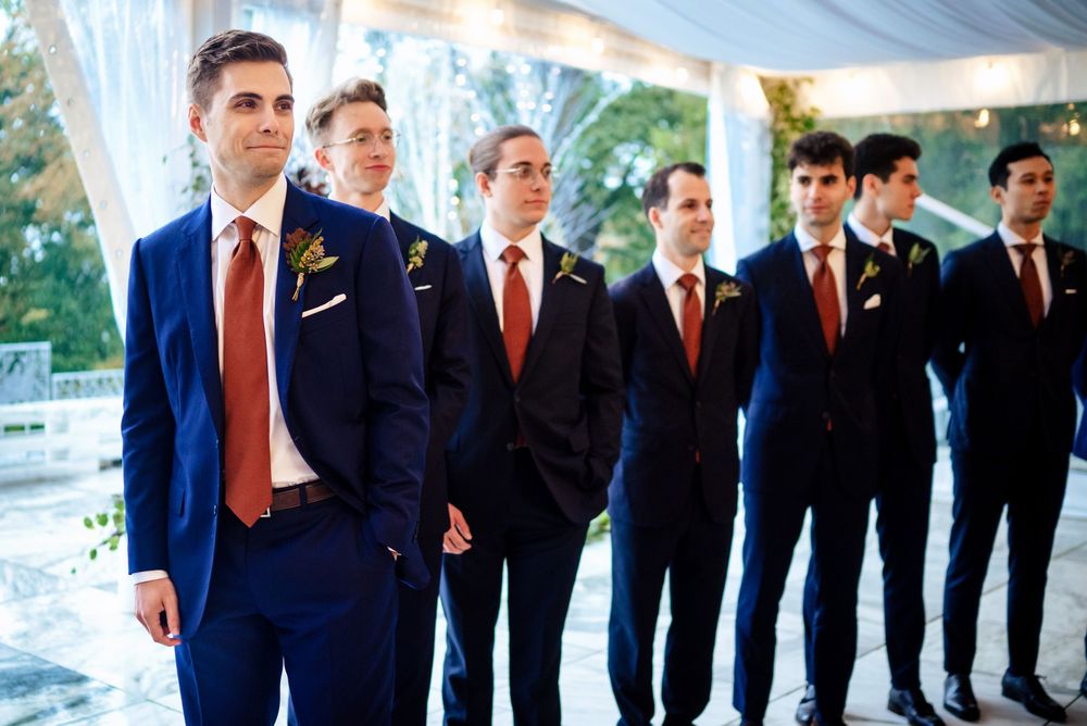 Groom and groomsmen in navy blue suits with red ties and boutonnieres standing at an outdoor wedding ceremony.