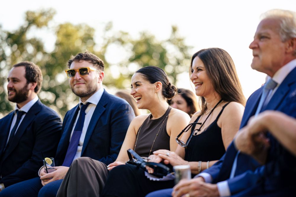 Smiling wedding guests in formal attire seated together enjoying an outdoor wedding ceremony.