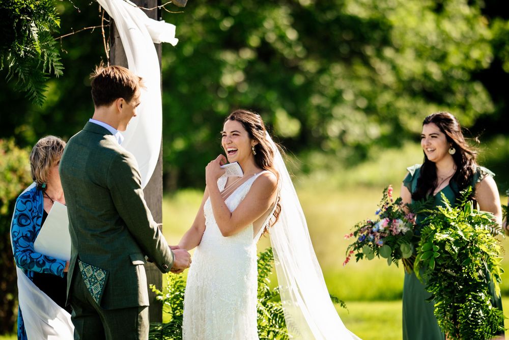Radiant bride laughs joyfully while holding grooms hands during outdoor garden wedding ceremony.