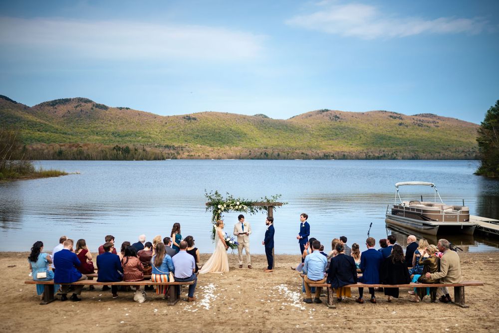 Intimate lakeside wedding ceremony with guests seated on wooden benches before scenic mountain backdrop.