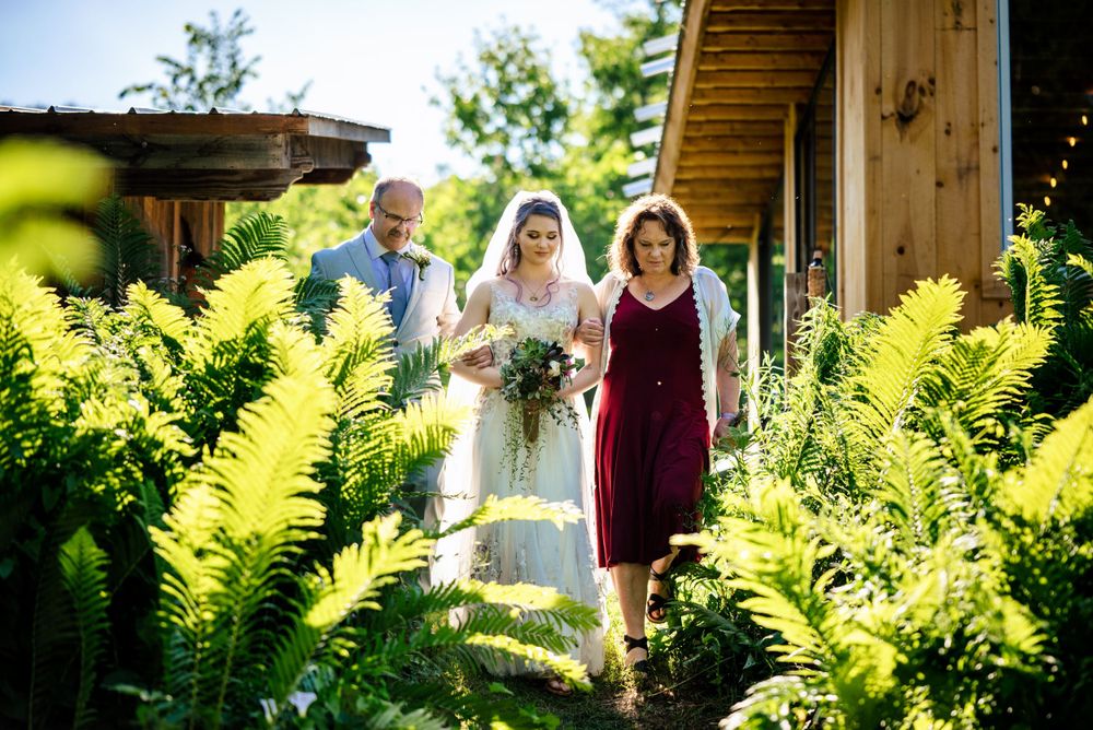Bride walks with parents through lush fern garden path toward rustic wooden wedding venue.