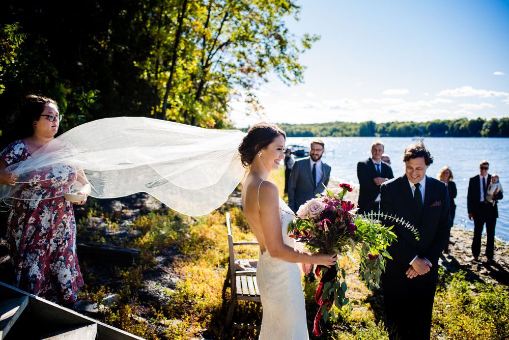 Bride's veil blows in wind as she stands at lakeside wedding ceremony on sunny autumn day.