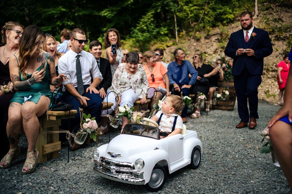 Adorable ring bearer rides miniature white vintage car down gravel wedding ceremony aisle.
