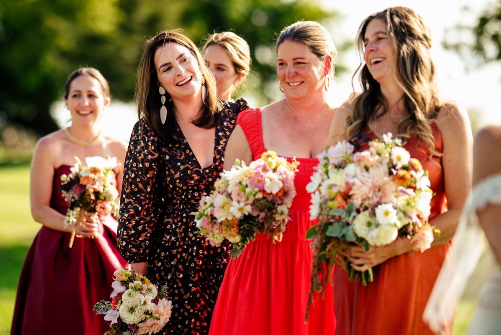 Five bridesmaids in colorful dresses holding floral bouquets laugh together outdoors at a sunny wedding celebration.