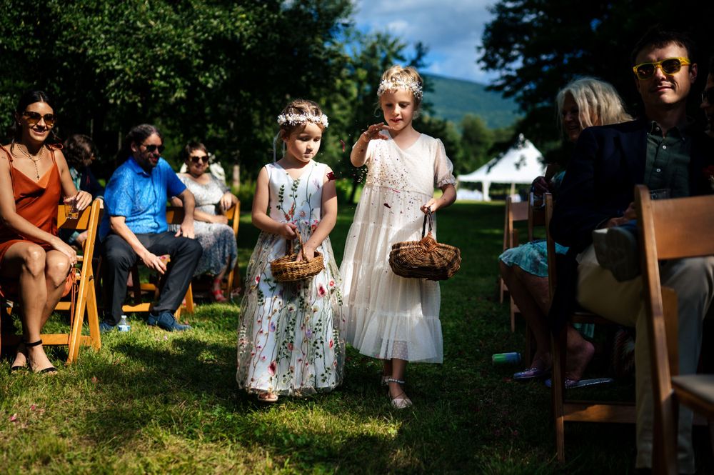 Two adorable flower girls in floral dresses walk down grassy outdoor wedding ceremony aisle.