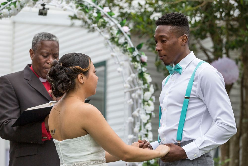 Groom in teal bow tie and suspenders holds bride's hands during outdoor wedding ceremony.