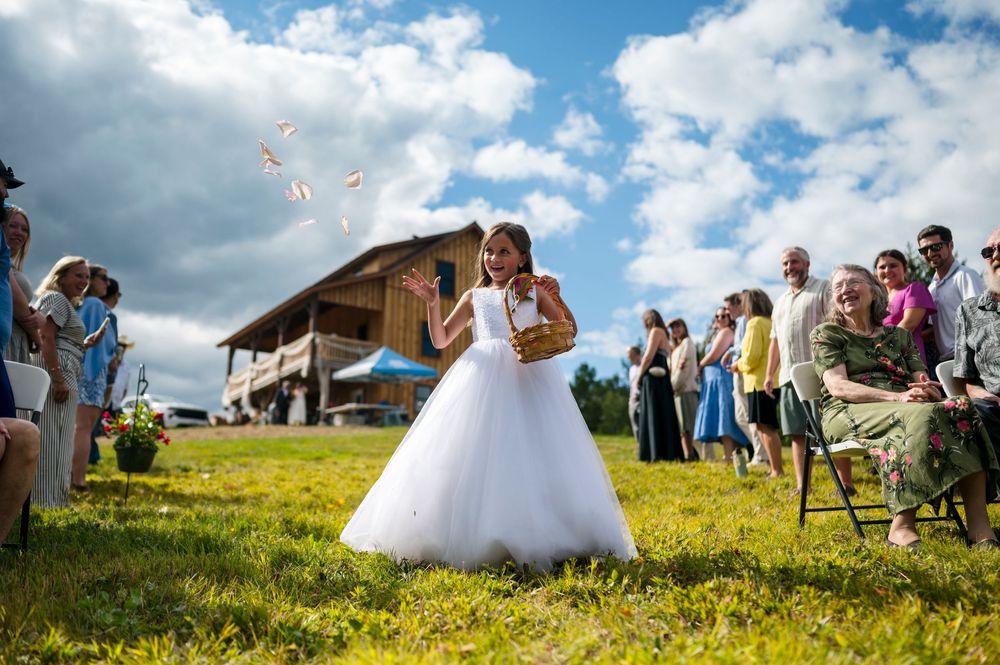 Young flower girl in white dress scattering petals at outdoor barn wedding ceremony on sunny day.