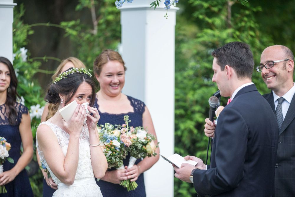 Emotional bride wipes tears while groom reads vows at outdoor wedding ceremony.
