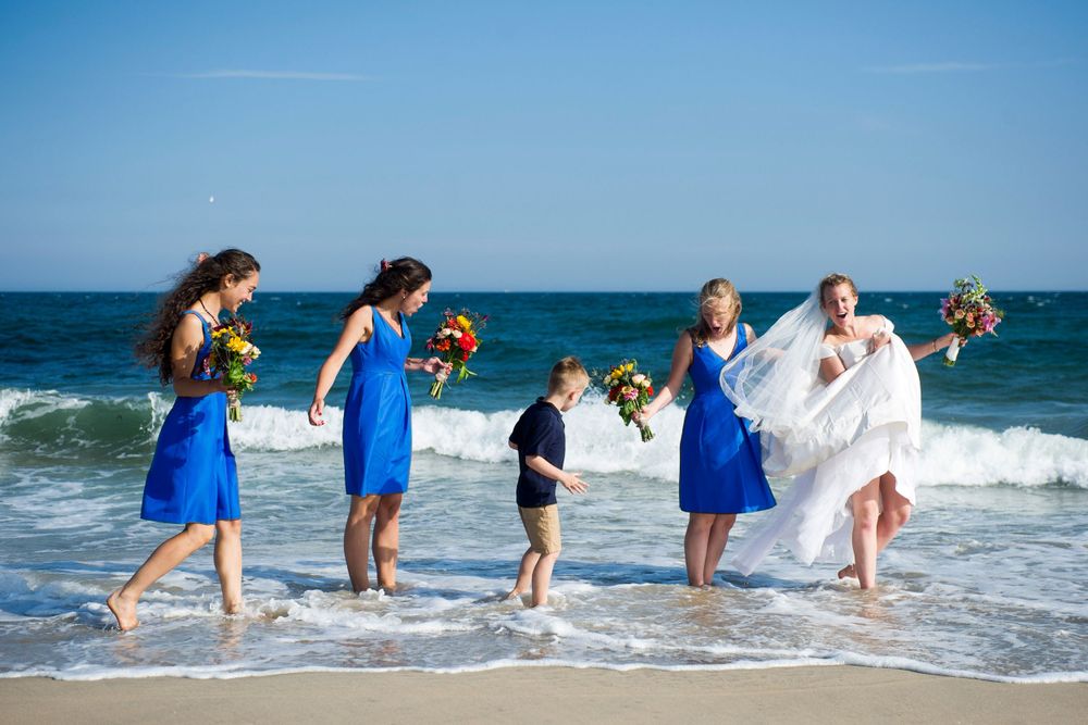 Bride and bridesmaids in blue dresses walk along ocean waves at beach wedding.