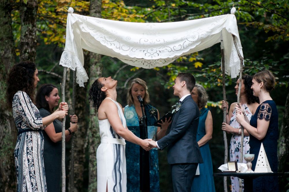 Couple laughing joyfully during outdoor Jewish wedding ceremony under white lace chuppah surrounded by wedding party.