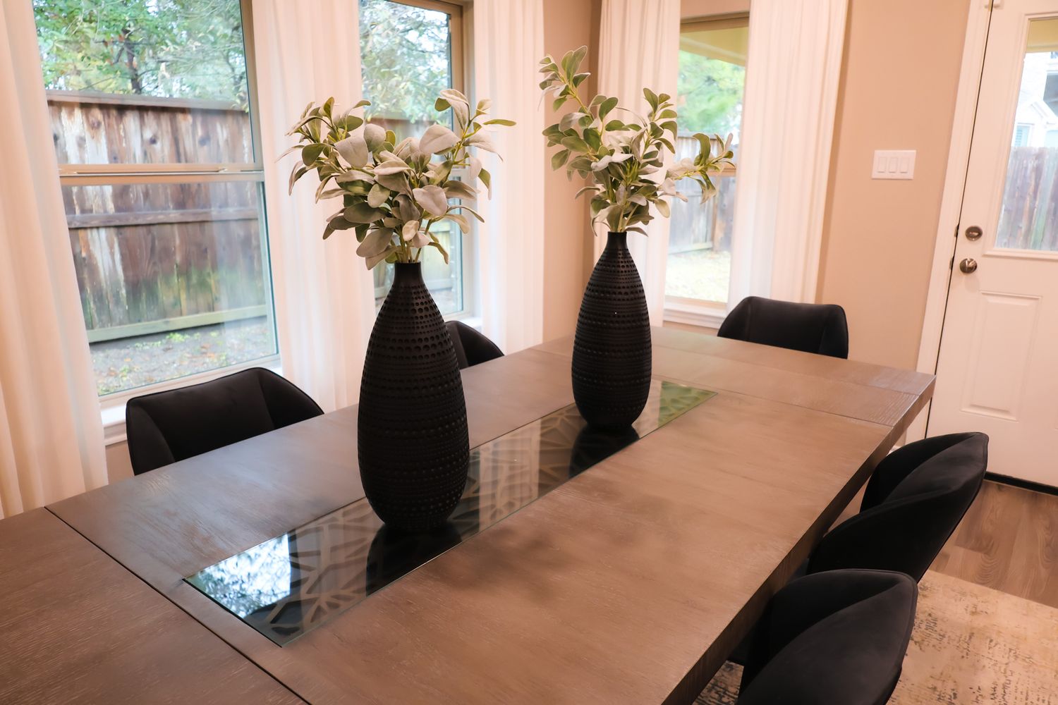 Modern dining table with black vases holding green plants against bright window backdrop.